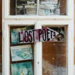 Vor einem verwitterten Fabrikfenster hängt ein handgemaltes Schild mit „LOST POETS“, das auf das L/OST POETRY Festival in Cottbus deutlich verweist.