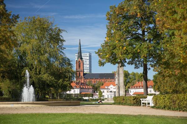 Im Park von Cottbus blickt man auf die Altstadt mit Kirche und Brunnen, passend zur Reihe „Sport in der City: Pilates mit Femaktiv“.