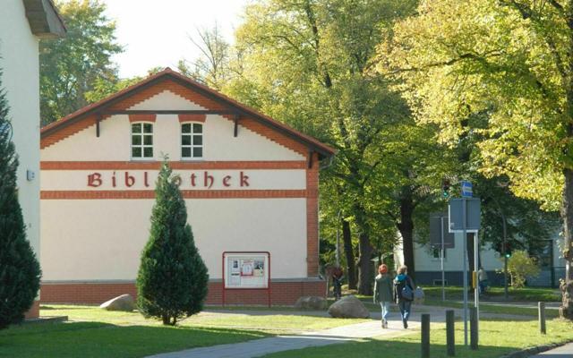 Ein Gebäude mit dem Schriftzug Bibliothek steht an einem Weg, davor gehen zwei Personen durch eine grüne Anlage.