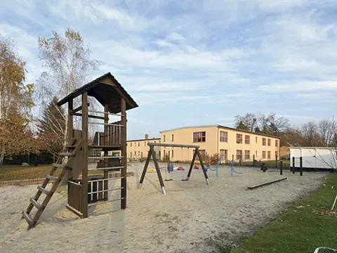 Ein Holz-Kletterturm und mehrere Schaukeln stehen auf einem sandigen Spielplatz vor einem hellen Kita-Gebäude.