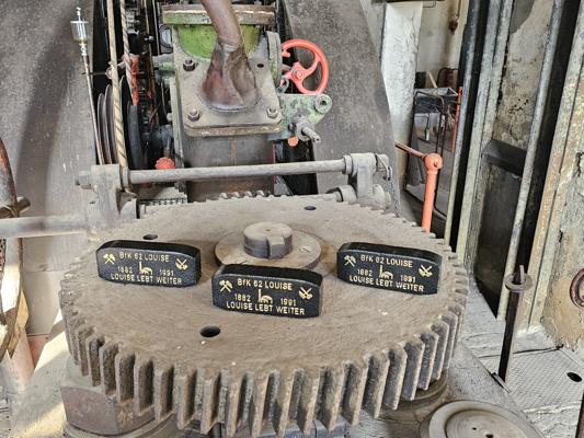 Eine verstaubte Industrieanlage mit großem Zahnrad, Hebeln und drei Schildern mit der Aufschrift »BFK 62 Louise«.