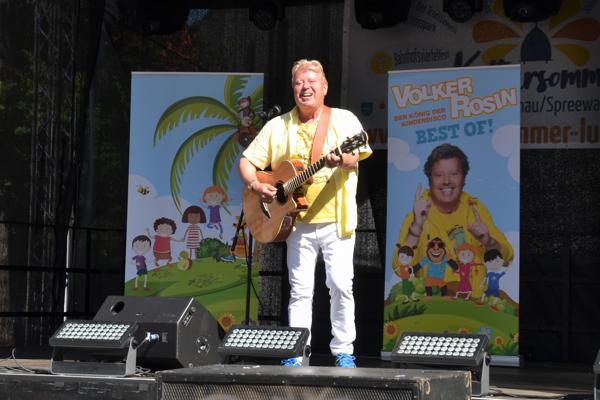 Volker Rosin singt und spielt Gitarre auf dem Spreewald- und Schützenfest in Lübbenau, vor bunten Kinderplakaten und hellen Bühnenlichtern.