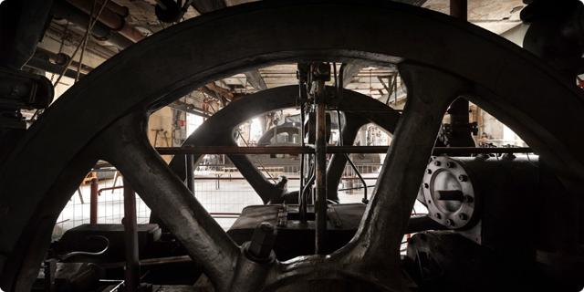 Große Schwungräder und Metallstreben einer historischen Dampfmaschine stehen in einem gedämpft beleuchteten Maschinenraum.