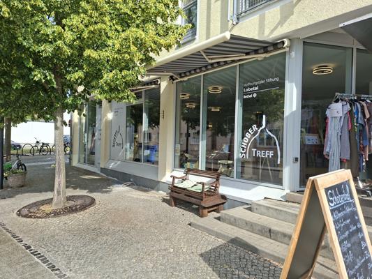 Vor dem Schieber-Treff in Cottbus steht eine Bank neben dem Gehweg, während vor dem Laden eine Tafel und Fahrräder den Eingangsbereich prägen.