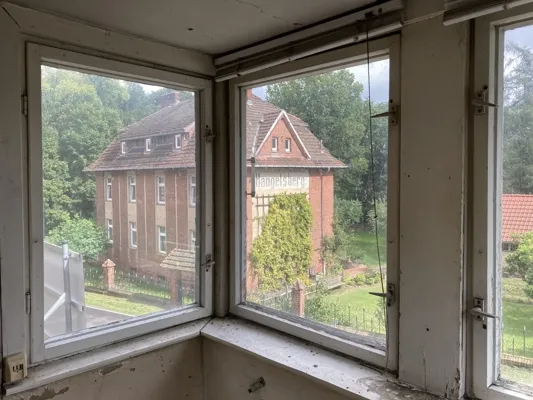 Blick aus einem alten Bahnhofsraum auf ein Backsteingebäude und grünes Umland in Hangelsberg, passend zur Ausstellung „Rasender Stillstand“.