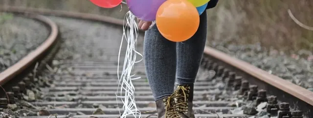 Eine Person steht auf Bahngleisen und hält bunte Luftballons mit langen weißen Schnüren in der Hand.