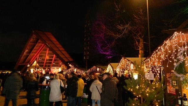 Am Festplatz stehen beleuchtete Holzbuden, davor versammelt sich eine Menschenmenge in winterlicher Nacht.