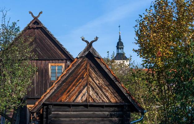 Traditionelle Holzhäuser mit steilen Dächern stehen unter blauem Himmel, dahinter ragt ein Kirchturm zwischen Bäumen auf.