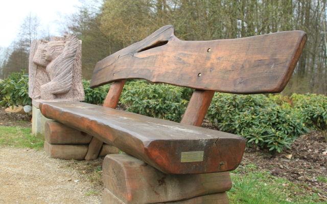 Eine massive Holzbank mit geschnitzter Lehne und Pferdekopf steht am Wegrand vor Hecken und Bäumen.