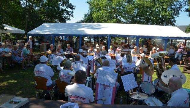 Ein Blasorchester spielt unter einem weißen Festzelt vor sitzendem Publikum, mit Tuben und Schlagzeug im Vordergrund.