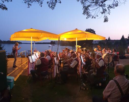 Am Ufer des Großen Müllroser Sees spielt ein Musikensemble in der Dämmerung unter gelben Sonnenschirmen.