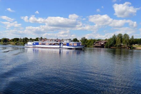 Ein weiß-blaues Ausflugsschiff fährt über ruhiges Wasser, am Ufer stehen Bäume und ein Steg unter einem wolkigen Himmel.