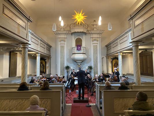 In der barocken Kirche spielt ein Orchester vor dem Altar, während ein Dirigent die Musiker unter dem Stern anleitet.