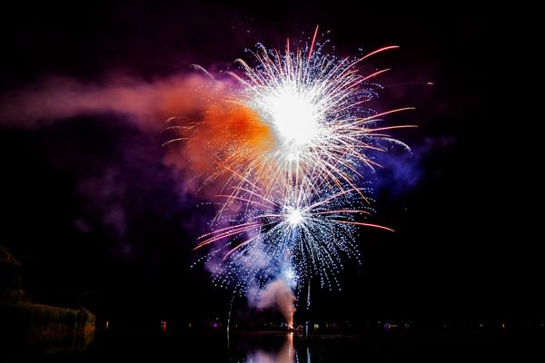 Mehrere Feuerwerkskörper explodieren über dem dunklen See, weiße, blaue und orangefarbene Lichter spiegeln sich im Wasser.