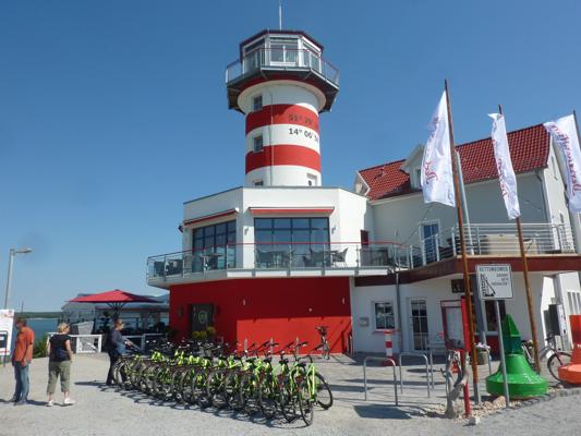 Vor einem rot-weiß gestreiften Leuchtturm stehen viele Fahrräder; daneben wehen weiße Fahnen am Gebäude am Hafen.