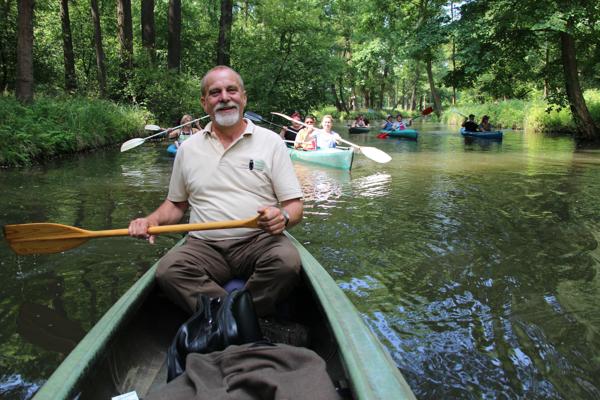 Ein älterer Mann sitzt mit Paddel im Kanu auf einem schmalen Fließ, hinter ihm fahren weitere Boote durch den dichten Wald.