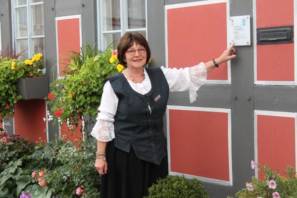 Eine Frau in historischer Kleidung lehnt an einer roten Haustür und zeigt auf ein Schild an der grauen Fassade neben Blumen.