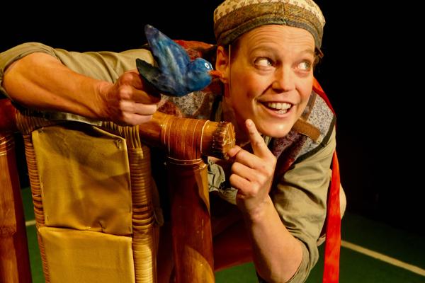 Eine lächelnde Theaterfigur im Kostüm beugt sich hinter eine Holzlehne und hält einen blauen Vogel in der Hand.