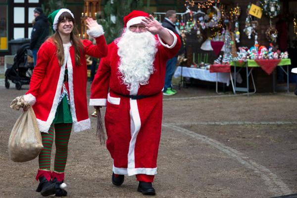 Ein Weihnachtsmann und eine Wichtelfigur winken auf einem Weihnachtsmarkt vor geschmückten Ständen und Lichterketten.