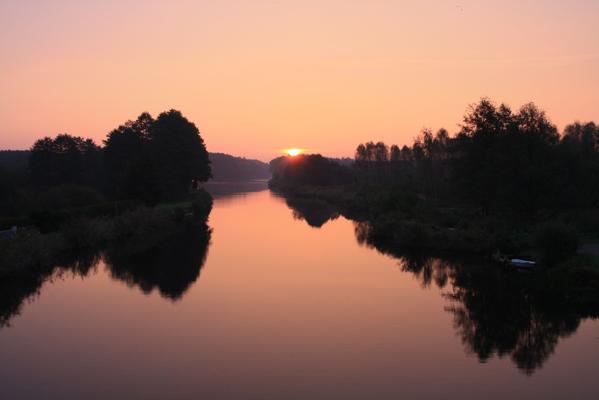Ein ruhiger Fluss spiegelt den orangefarbenen Himmel bei Sonnenuntergang, flankiert von dunklen Baumreihen.