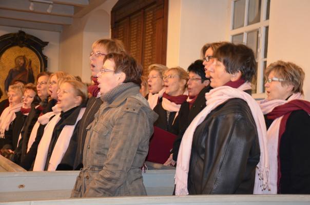 Mehrere Frauen eines Chors singen in einem hellen Kirchenraum, viele tragen Schals und Winterjacken.
