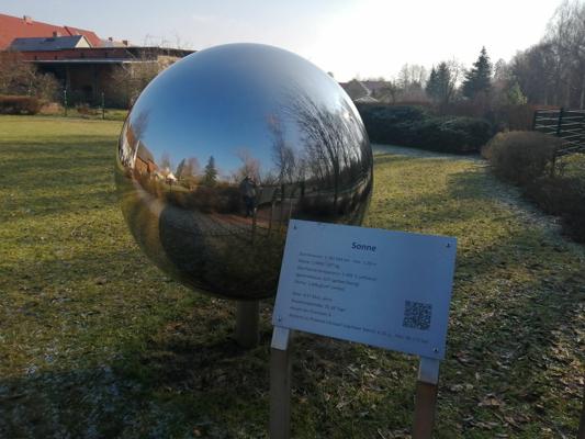 Eine große Spiegelkugel steht auf einer Wiese, daneben erklärt eine Tafel mit der Überschrift Sonne das Modell.
