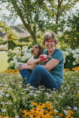 Eine lachende Frau mit roter Brille hält einen Hund auf dem Schoß und sitzt inmitten von Blumen im Garten.