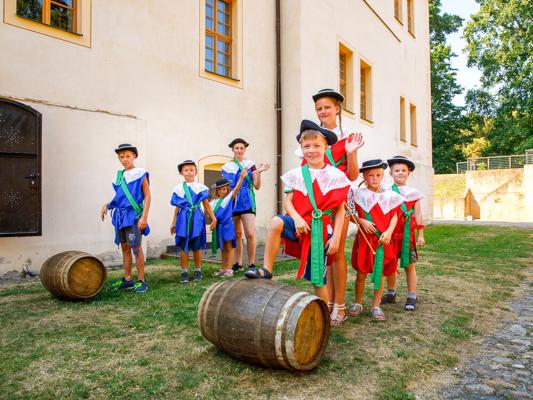 Kinder in roten und blauen Musketierkostümen stehen mit einer Begleiterin vor einer hellen Schlossmauer und zwei Holzfässern.