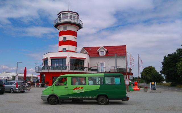Ein grüner Kleinbus steht vor einem rot-weiß gestreiften Leuchtturmgebäude mit rotem Dach und bewölktem Himmel.