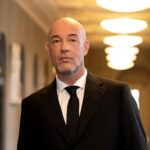 Ein kahlköpfiger Mann im schwarzen Anzug mit weißem Hemd und schwarzer Krawatte steht in einem hell beleuchteten Flur.
