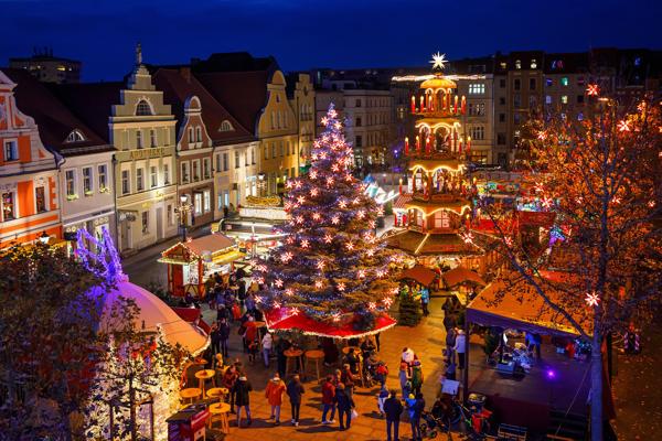 Im festlich beleuchteten Cottbuser Weihnachtsmarkt stehen ein geschmückter Baum und eine mehrstöckige Weihnachtspyramide.