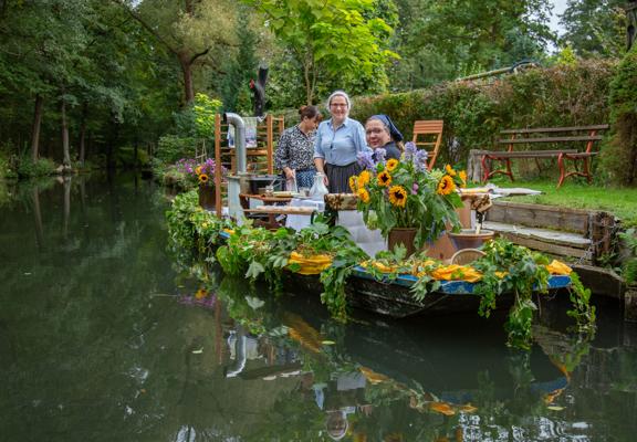 Drei Personen stehen auf einem bunt geschmückten Kahn mit Blumen und Sonnenblumen, der ruhig auf einem schmalen Kanal fährt.
