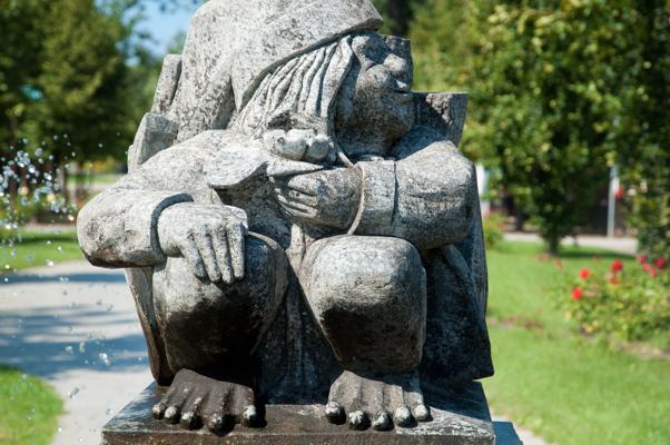 Die sitzende Steinfigur mit Kapuze und gesenktem Blick steht an einem Weg im grünen Park, umgeben von Rasen und Blumen.