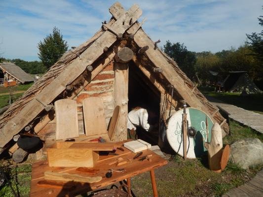 Vor einer strohgedeckten Lehmhütte stehen Holzwerkzeuge, Bretter und ein runder Schild im Freilichtmuseum.