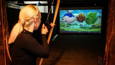 Eine blonde Frau zielt mit dem Bogen auf eine Leinwand mit Drachen- und Landschaftsprojektion in einem dunklen Raum.