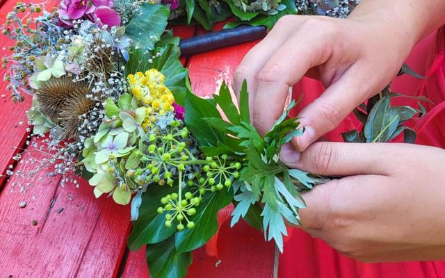 Hände binden einen Herbstkranz aus grünen Blättern, gelben Blüten und getrockneten Pflanzen auf einem roten Holztisch.