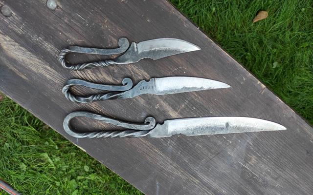 Drei handgeschmiedete Messer mit gedrehten Griffen liegen nebeneinander auf einer dunklen Holzbank im Gras.
