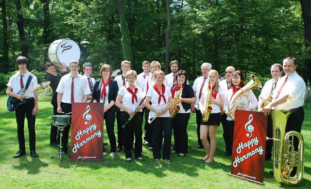 Eine Musikgruppe in weißen Hemden und roten Halstüchern steht auf einer Wiese vor Bäumen, daneben „Happy Harmony“-Banner.