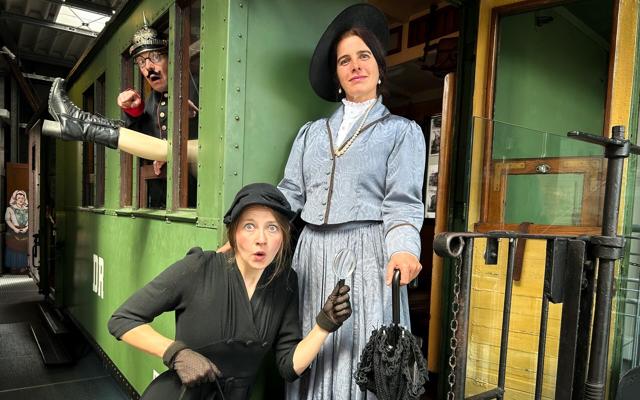 In einem grünen historischen Bahnwagen stehen zwei verkleidete Darsteller, während ein Mann aus dem Fenster schaut.
