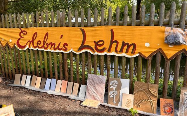 Ein Holzschild mit der Aufschrift „Elebni's Lehm“ hängt am Lattenzaun; darunter stehen Lehmputzproben in Plattenform.