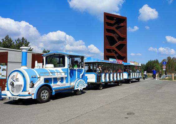 Eine blau-weiße Bimmelbahn mit Waggons steht vor einem roten Aussichtsturm, daneben gehen Besucher.