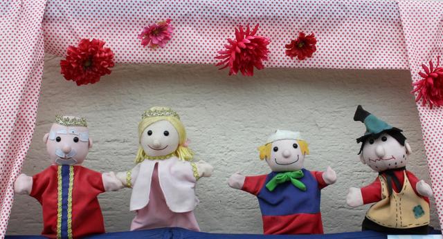Vier Kasperpuppen stehen vor einem gepunkteten Vorhang, darüber hängen rote und pinke Papierblumen.