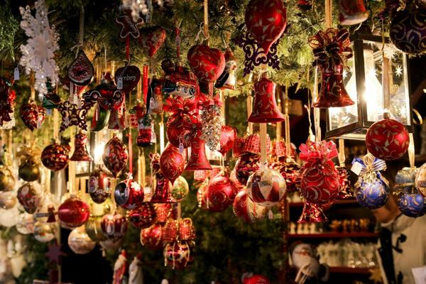 Zahlreiche rote und goldene Glasornamente hängen dicht an einem dekorierten Marktstand zwischen Tannengrün und warmem Licht.