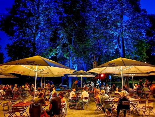 Gäste sitzen im Biergarten unter gelben Sonnenschirmen, während Livemusik vor blau angestrahlten Bäumen erklingt.