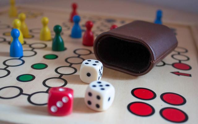 Würfel, bunte Spielfiguren und ein Lederbecher liegen auf einem Spielbrett mit markierten Feldern.