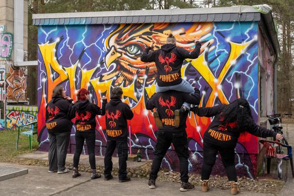 Mehrere Personen in schwarzen Phoenix-Hoodies posieren vor einer bunten Graffitiwand mit großem Auge und Blitzmotiven.