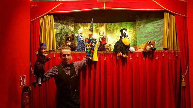 Vor einem roten Vorhang steht ein Spieler mit Handpuppen, hinter ihm sind mehrere Kasperfiguren auf der Bühne zu sehen.