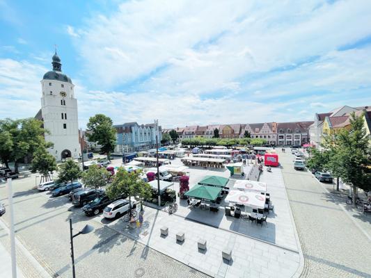 Auf dem Marktplatz stehen mehrere Marktstände, daneben ein hoher Stadtturm und historische Häuser unter blauem Himmel.
