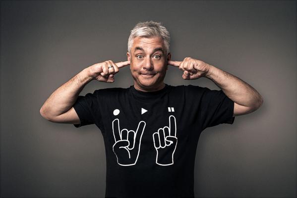 Ein grauhaariger Mann hält sich mit beiden Zeigefingern die Ohren zu und trägt ein schwarzes T-Shirt mit Rockhand-Symbolen.