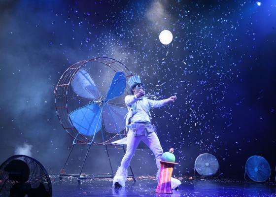 Eine Performerfigur steht vor einem großen Ventilator, während weiße Partikel wie Schnee über die dunkle Bühne wirbeln.